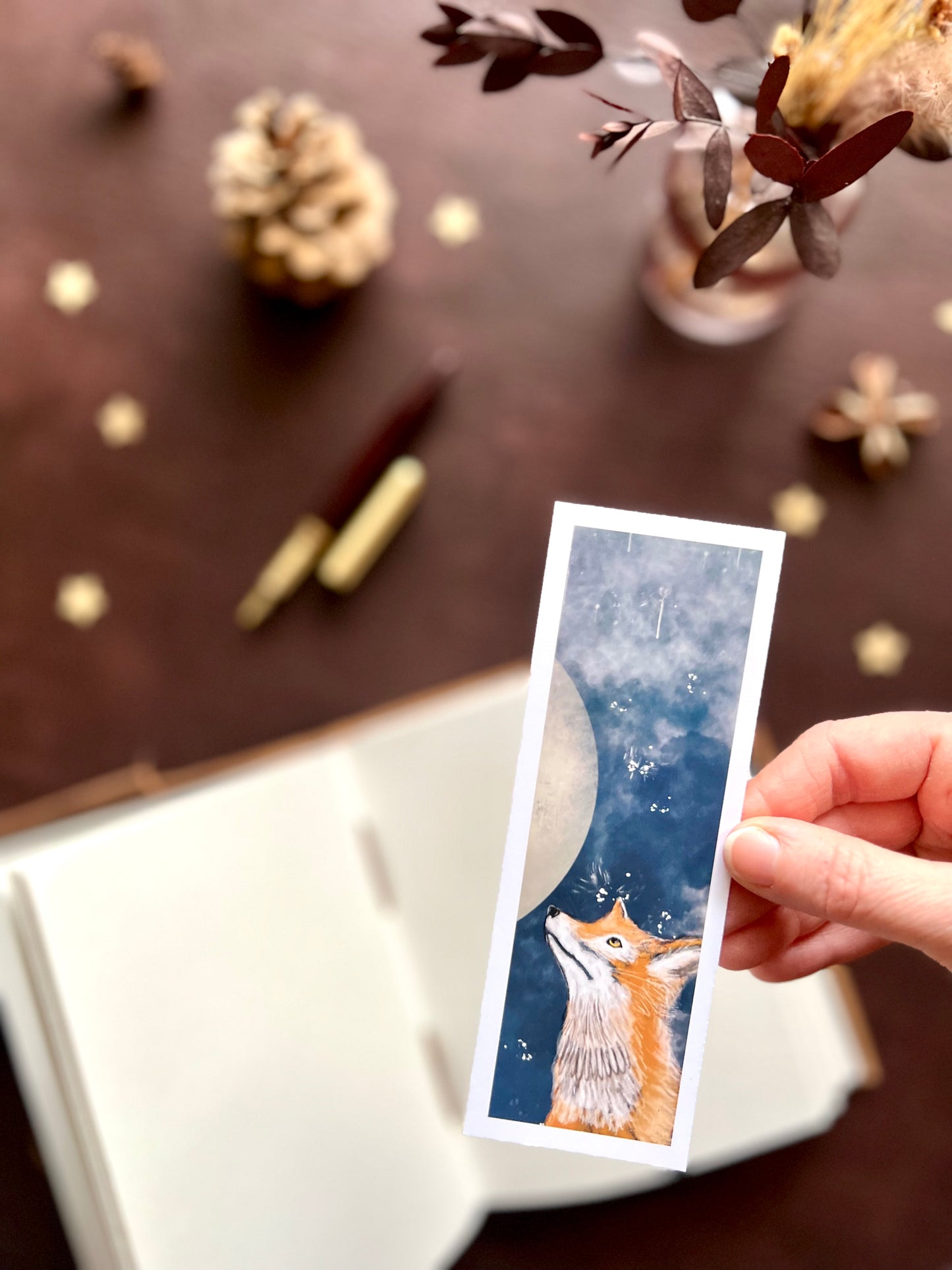 Hand holding a card with a fox design against a blurred decorative background. Dark academia and nature theme.