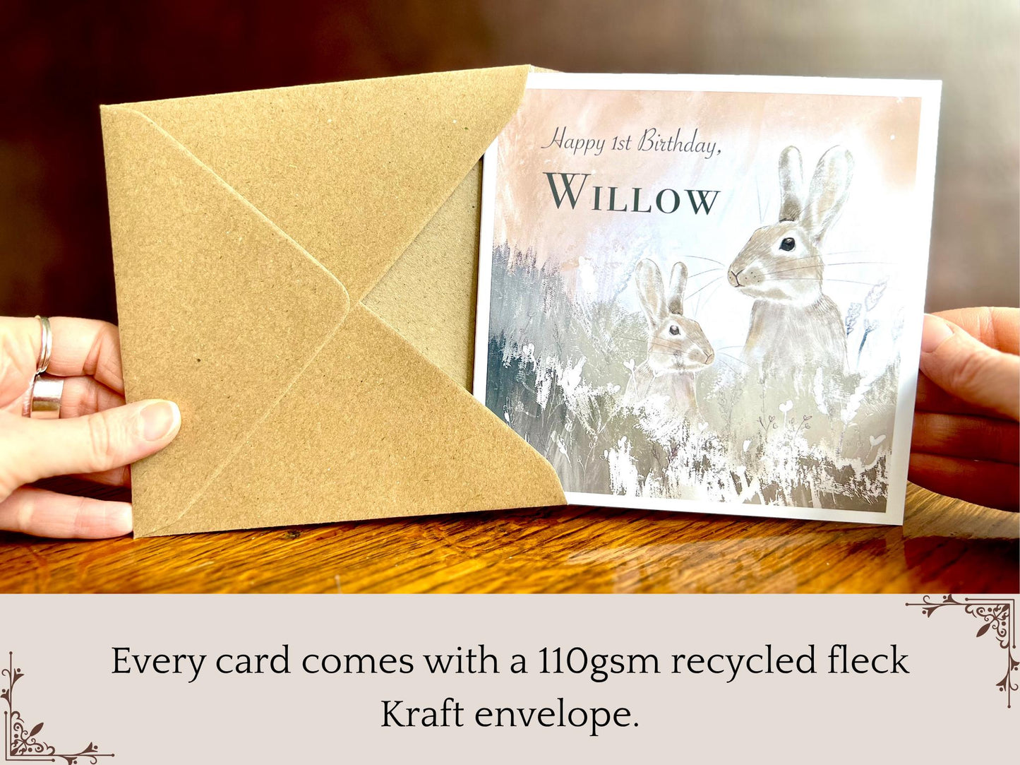 Hand holding a handmade customisable rabbit birthday card being removed from a kraft envelope, shown upright towards the camera. The card features a silvery grey floral illustration with a pale pink evening sky background, and text highlighting that every card comes with a 110gsm recycled fleck Kraft envelope.