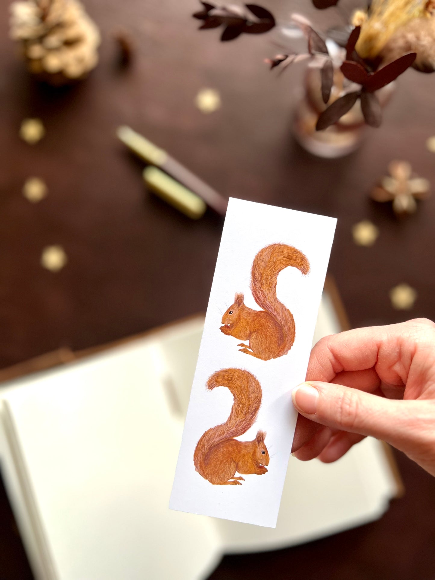 Hand holding a red squirrel bookmark featuring two illustrated squirrels, with softly blurred pine cones and flowers in a nature inspired background.