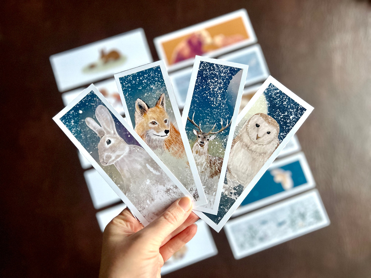Hand holding four illustrated animal bookmarks with rabbit, fox, owl, and deer designs, with the rest of the 16 pack set blurred in the background. Perfect for whimsical, rustic, or woodland weddings, and as wedding stationery or gifts.
