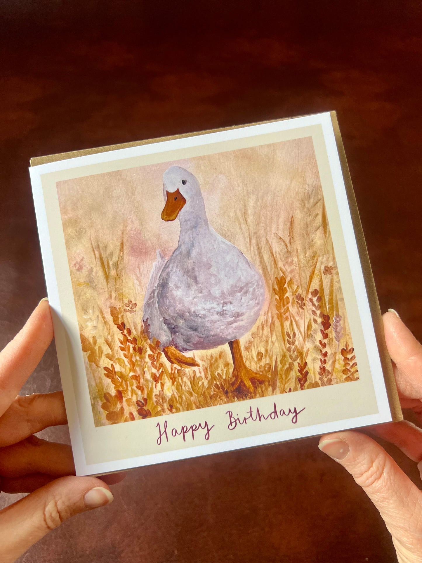 Hands holding a square Duck in the Meadow birthday card with matching envelope, featuring a characterful white duck in orange flowers with deep red handwritten Happy Birthday text below the duck painting