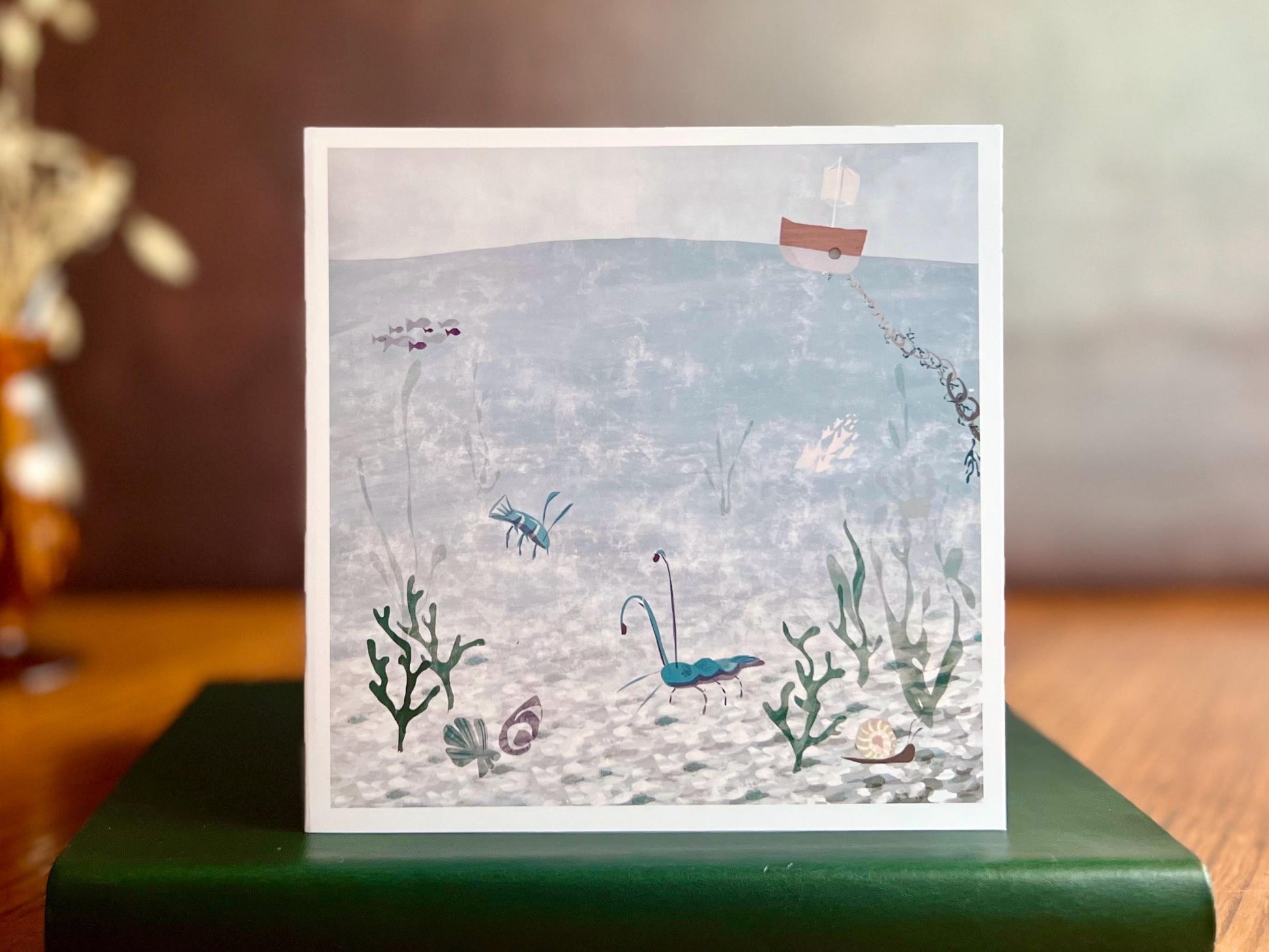 Artisan illustrated greeting card featuring a blue lobster and blue prawn in a crystal blue underwater coastal scene with a small boat above the water, displayed standing on a book. Joyful summery coastal art greeting card suitable for birthdays or keeping in touch.