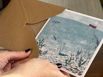 Artisan illustrated greeting card featuring a blue lobster and blue prawn in a crystal blue underwater sea scene with a small boat above the water, shown in close up being removed from a recycled fleck kraft envelope. Joyful summery sealife art greeting card suitable for birthdays or keeping in touch.