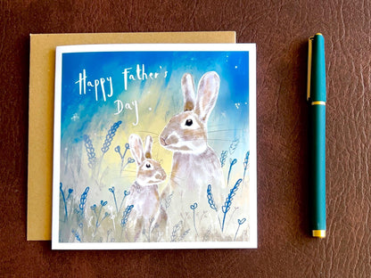 Artisan Father’s Day greeting card laid flat on a leatherette tabletop surface, featuring an illustrated father rabbit and young rabbit sitting in foliage under a starry blue and green night sky. The front reads “Happy Father’s Day”. Premium wildlife-inspired greeting card with nature-themed design.