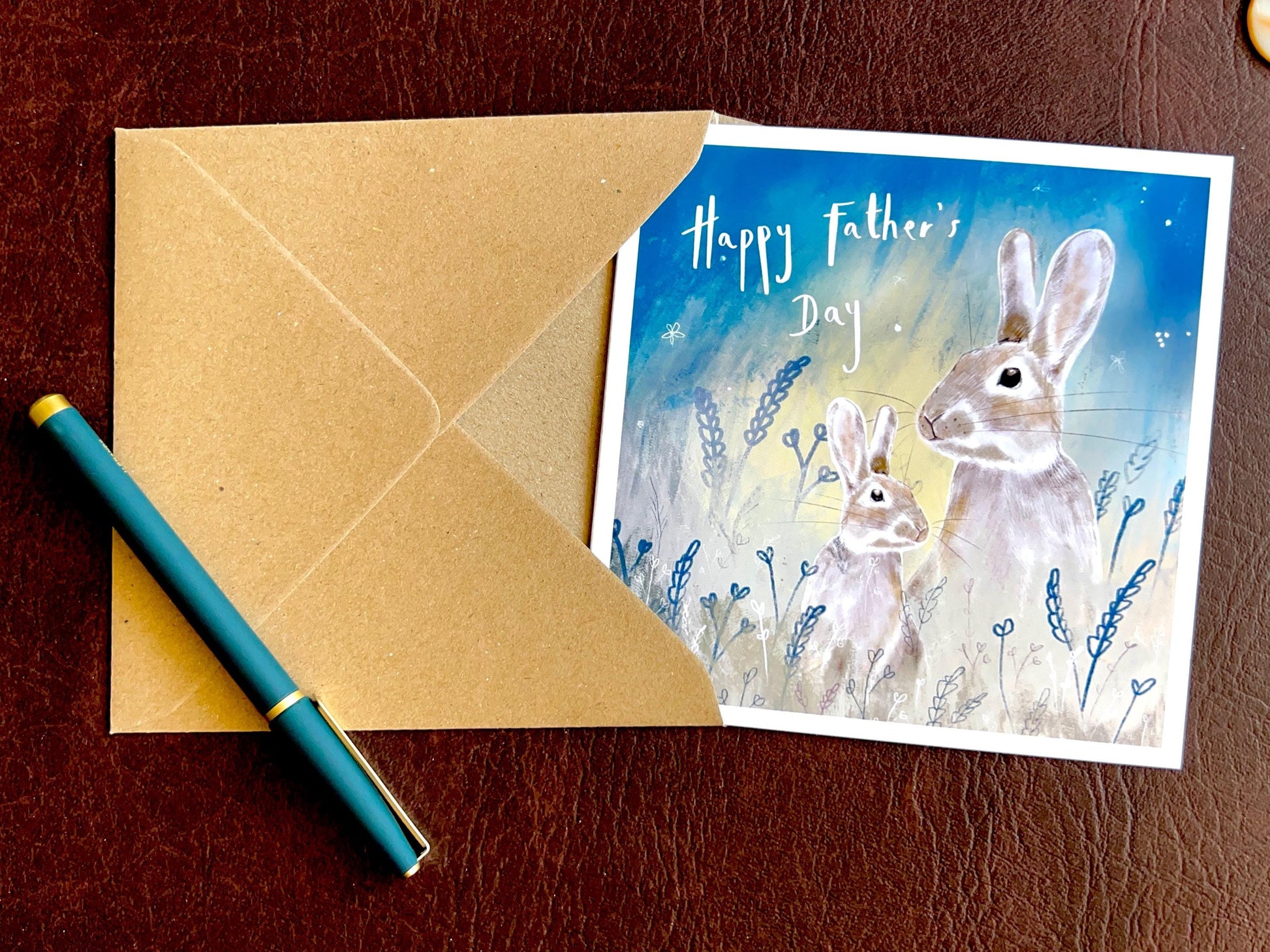Artisan Father’s Day greeting card featuring an illustrated father rabbit and young rabbit under a starry night sky, laid flat on a leatherette tabletop with a recycled fleck kraft envelope and pen. Premium wildlife-themed greeting card flat lay showcasing design detail and presentation.