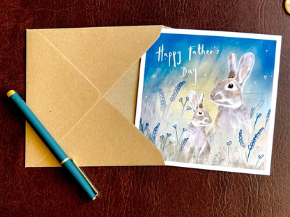 Artisan Father’s Day greeting card featuring an illustrated father rabbit and young rabbit under a starry night sky, laid flat on a leatherette tabletop with a recycled fleck kraft envelope and pen. Premium wildlife-themed greeting card flat lay showcasing design detail and presentation.