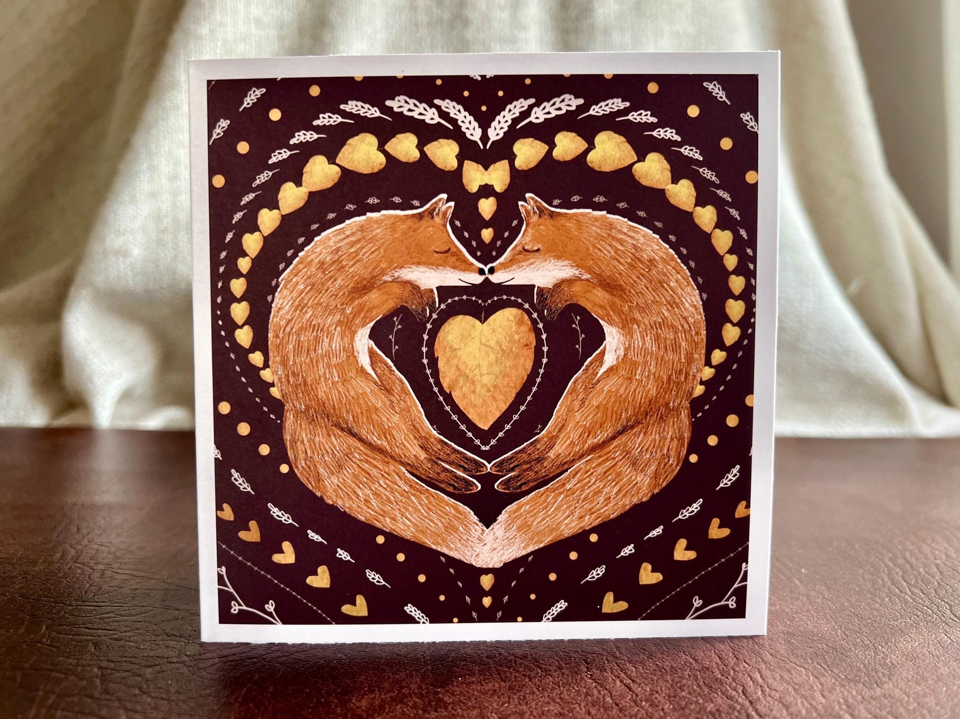 Artisan illustrated greeting card featuring two foxes sleeping nose-to-nose with their bodies curled into a heart shape, surrounded by a symmetrical leaf and heart pattern on a deep maroon red background. Romantic love-themed card suitable for Valentine’s Day or anniversaries, displayed standing upright on a tabletop.