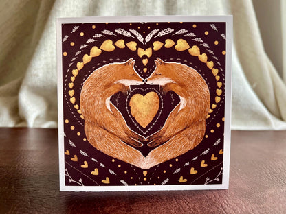 Artisan illustrated greeting card featuring two foxes sleeping nose-to-nose with their bodies curled into a heart shape, surrounded by a symmetrical leaf and heart pattern on a deep maroon red background. Romantic love-themed card suitable for Valentine’s Day or anniversaries, displayed standing upright on a tabletop.