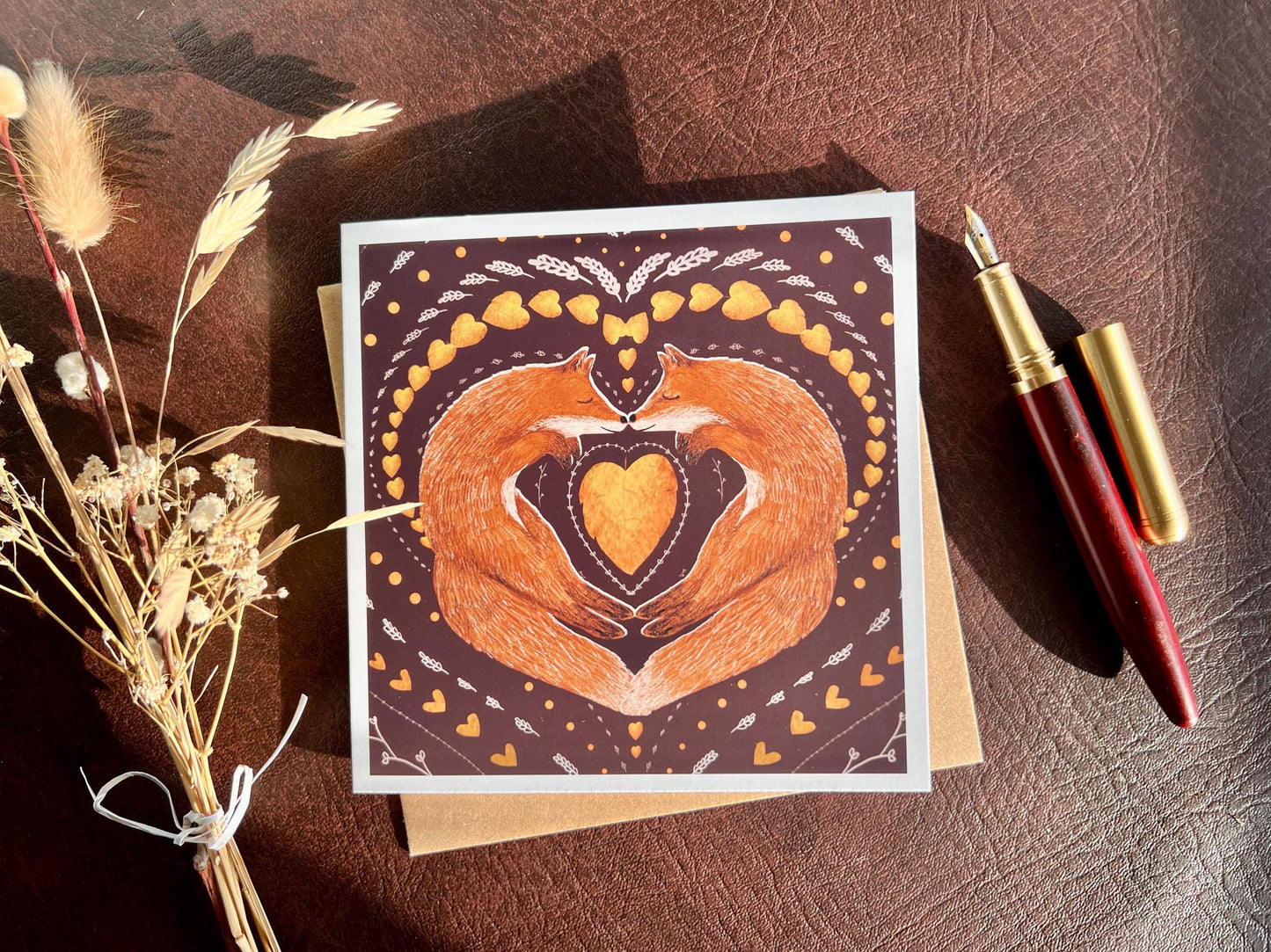 Artisan illustrated greeting card featuring two foxes sleeping nose-to-nose with their bodies curled into a heart shape, surrounded by a symmetrical leaf and heart pattern on a deep maroon red background. Romantic love-themed card suitable for Valentine’s Day or anniversaries, styled in a flat lay with a fountain pen and dried flowers.