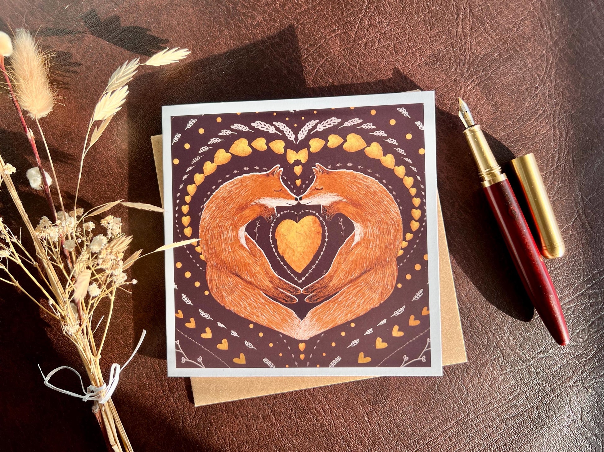 Artisan illustrated greeting card featuring two foxes sleeping nose-to-nose with their bodies curled into a heart shape, surrounded by a symmetrical leaf and heart pattern on a deep maroon red background. Romantic love-themed card suitable for Valentine’s Day or anniversaries, styled in a flat lay with a fountain pen and dried flowers.