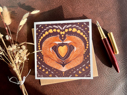 Artisan illustrated greeting card featuring two foxes sleeping nose-to-nose with their bodies curled into a heart shape, surrounded by a symmetrical leaf and heart pattern on a deep maroon red background. Romantic love-themed card suitable for Valentine’s Day or anniversaries, styled in a flat lay with a fountain pen and dried flowers.