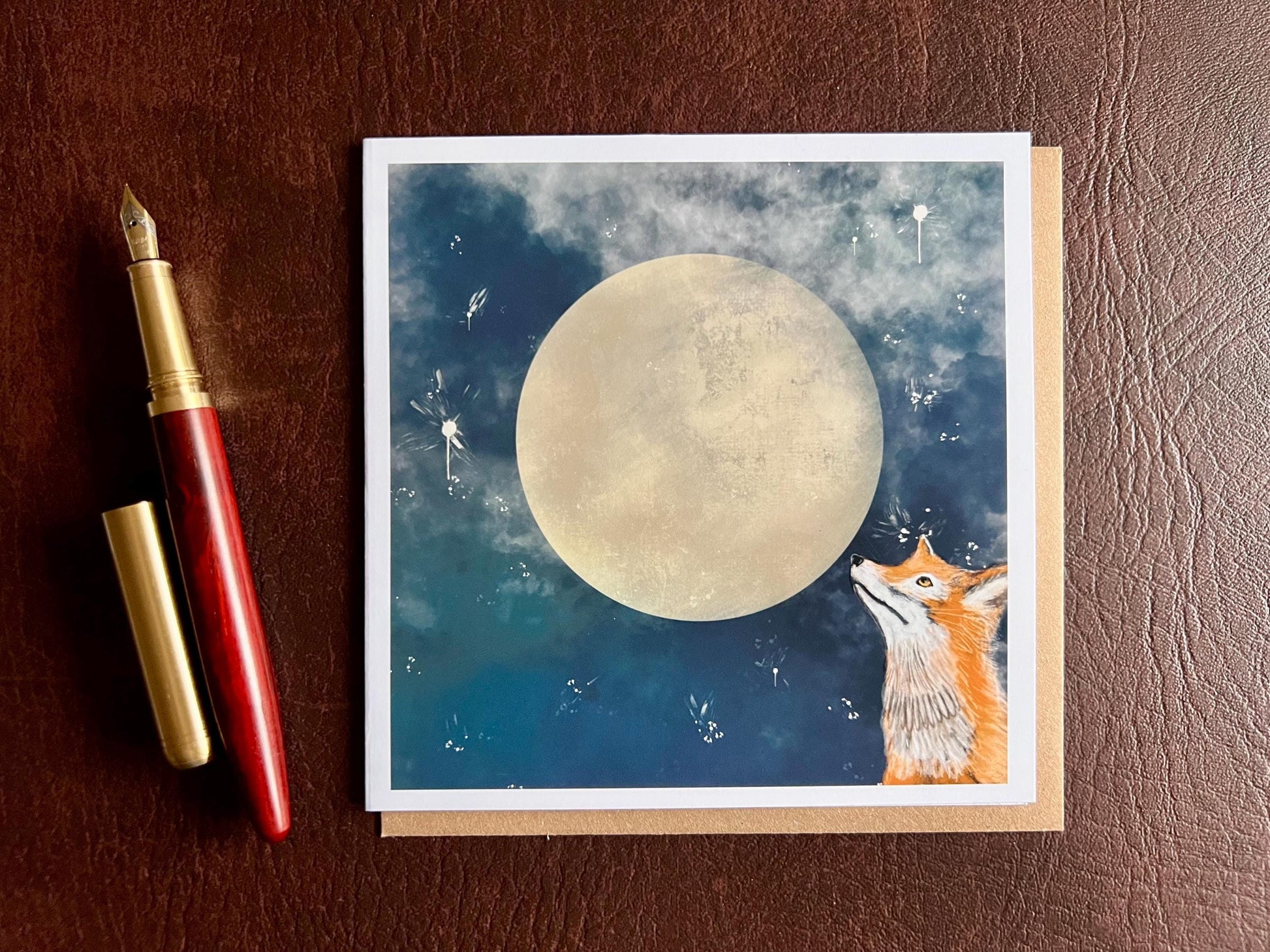Artisan illustrated greeting card featuring a fox looking up at a full moon in a blue starry night sky, laid flat on top of a recycled fleck kraft envelope next to a fountain pen in a styled flat lay composition showcasing premium wildlife artwork and handcrafted design.