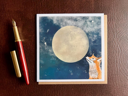 Artisan illustrated greeting card featuring a fox looking up at a full moon in a blue starry night sky, laid flat on top of a recycled fleck kraft envelope next to a fountain pen in a styled flat lay composition showcasing premium wildlife artwork and handcrafted design.