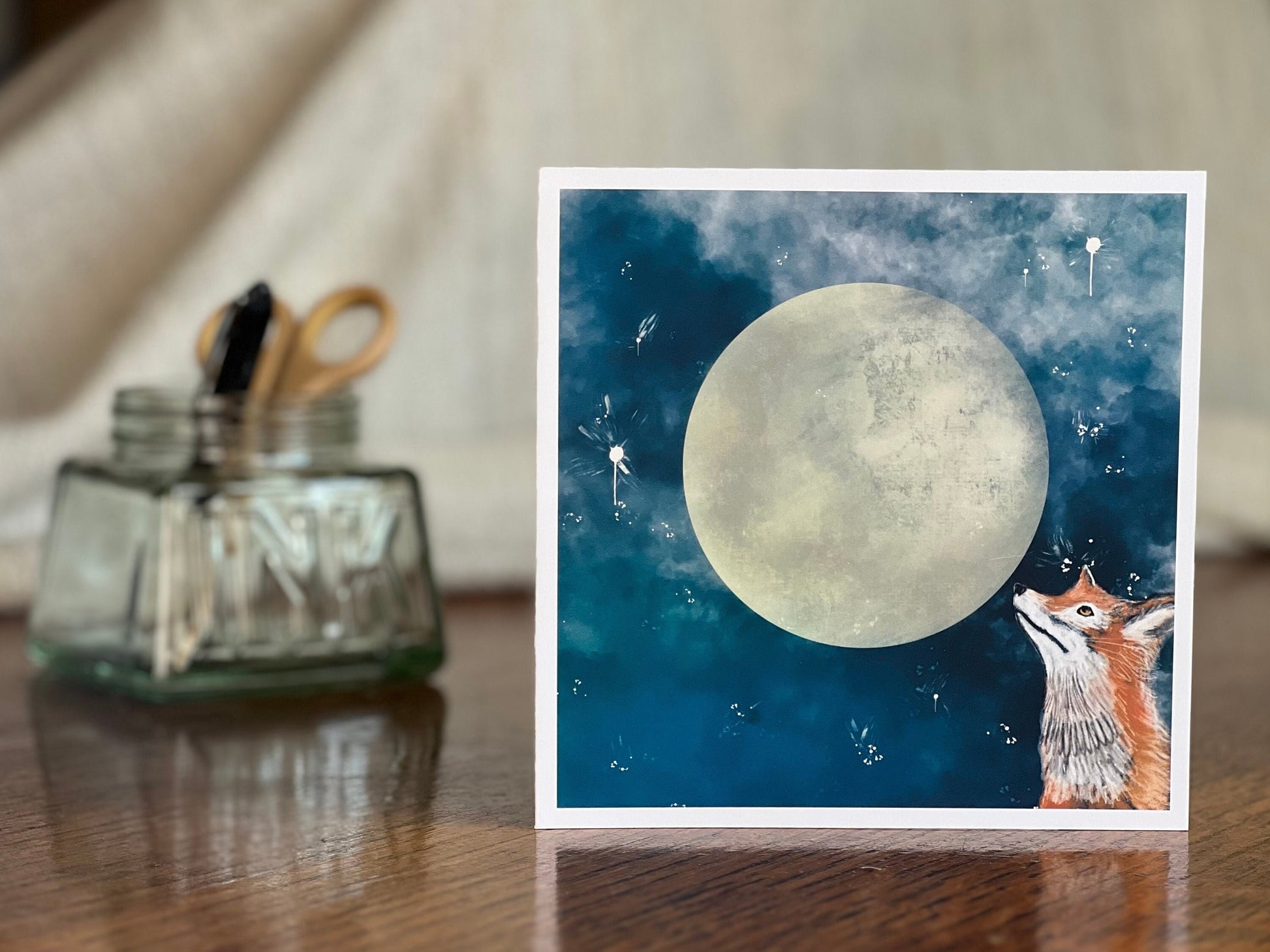 Artisan illustrated greeting card featuring a fox looking up at a full moon in a blue starry night sky, displayed standing upright on a tabletop. Premium wildlife art greeting card showcasing detailed nighttime illustration and handcrafted design.