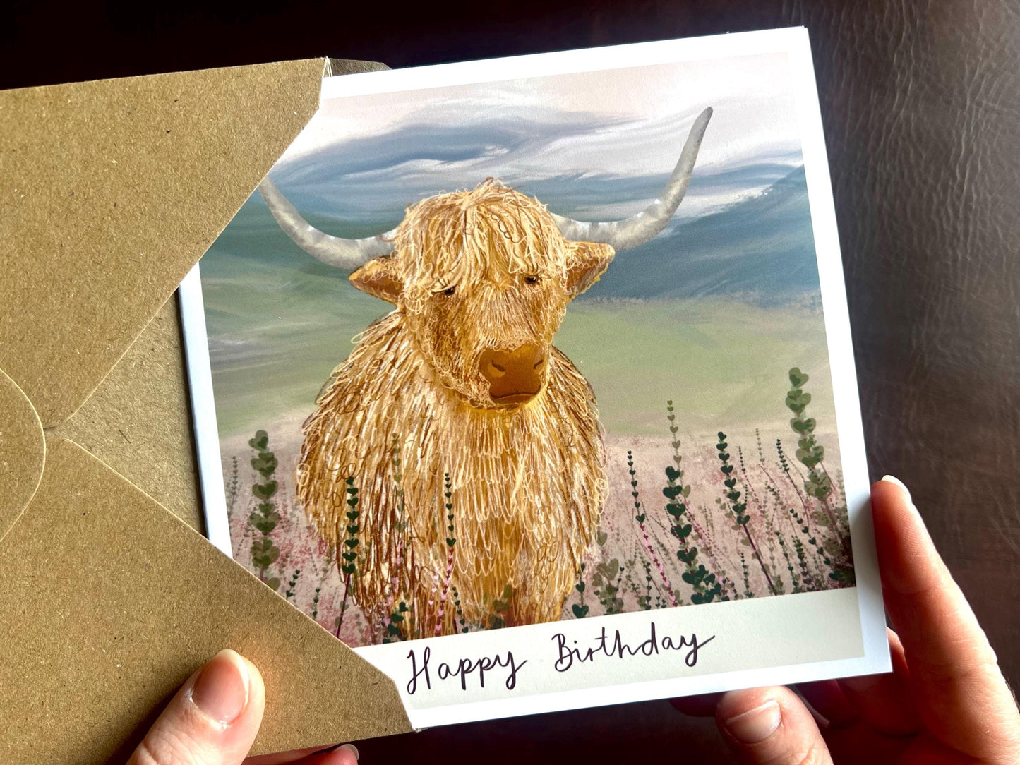 Artisan illustrated birthday greeting card featuring a highland cow standing amongst heather in a tranquil countryside landscape. The card shows the text “Happy Birthday” under the illustration and is being removed from a recycled fleck kraft envelope and held in hands.