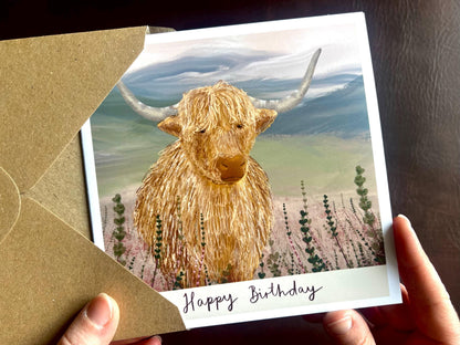 Artisan illustrated birthday greeting card featuring a highland cow standing amongst heather in a tranquil countryside landscape. The card shows the text “Happy Birthday” under the illustration and is being removed from a recycled fleck kraft envelope and held in hands.