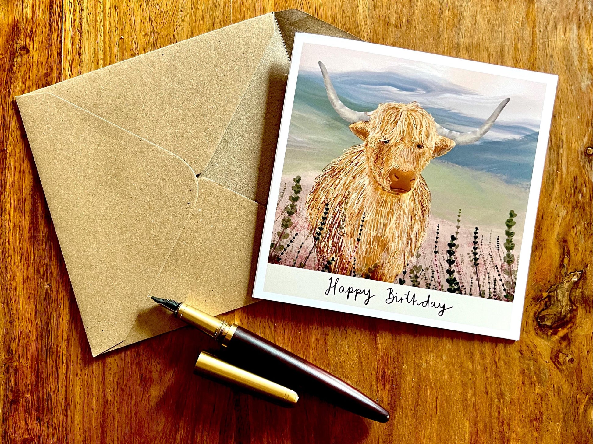 Artisan illustrated birthday greeting card featuring a highland cow standing amongst heather in a tranquil countryside landscape, laid flat on a tabletop with a recycled fleck kraft envelope. The design includes the text “Happy Birthday” beneath the illustration.