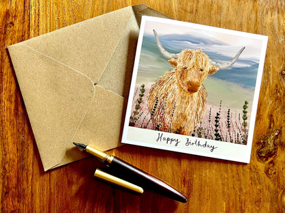 Artisan illustrated birthday greeting card featuring a highland cow standing amongst heather in a tranquil countryside landscape, laid flat on a tabletop with a recycled fleck kraft envelope. The design includes the text “Happy Birthday” beneath the illustration.