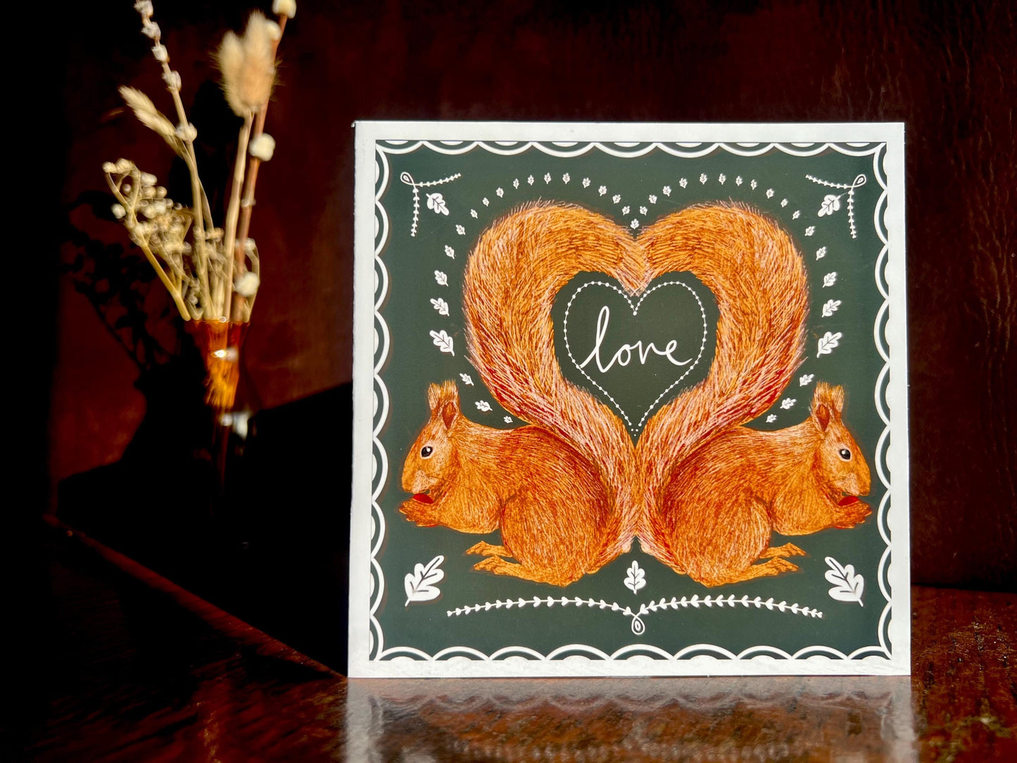 Artisan illustrated greeting card featuring two red squirrels forming a heart shape with their tails and the word “love” inside, displayed standing upright on a tabletop against a dark background. Romantic Valentine’s Day and anniversary card with dark green design and white scalloped border.