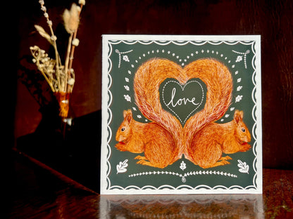 Artisan illustrated greeting card featuring two red squirrels forming a heart shape with their tails and the word “love” inside, displayed standing upright on a tabletop against a dark background. Romantic Valentine’s Day and anniversary card with dark green design and white scalloped border.