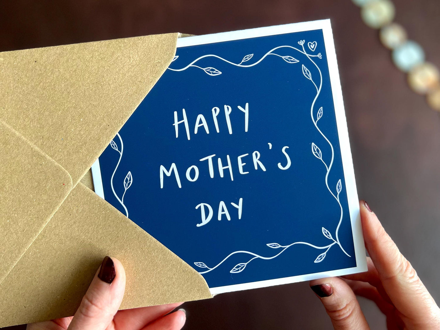 Artisan Mother’s Day greeting card with a dark navy blue background and simple leaf border, shown held in hands without personalisation. The centre reads “Happy Mother’s Day” with space for optional custom name text such as Mum, grandma, or step-mum.