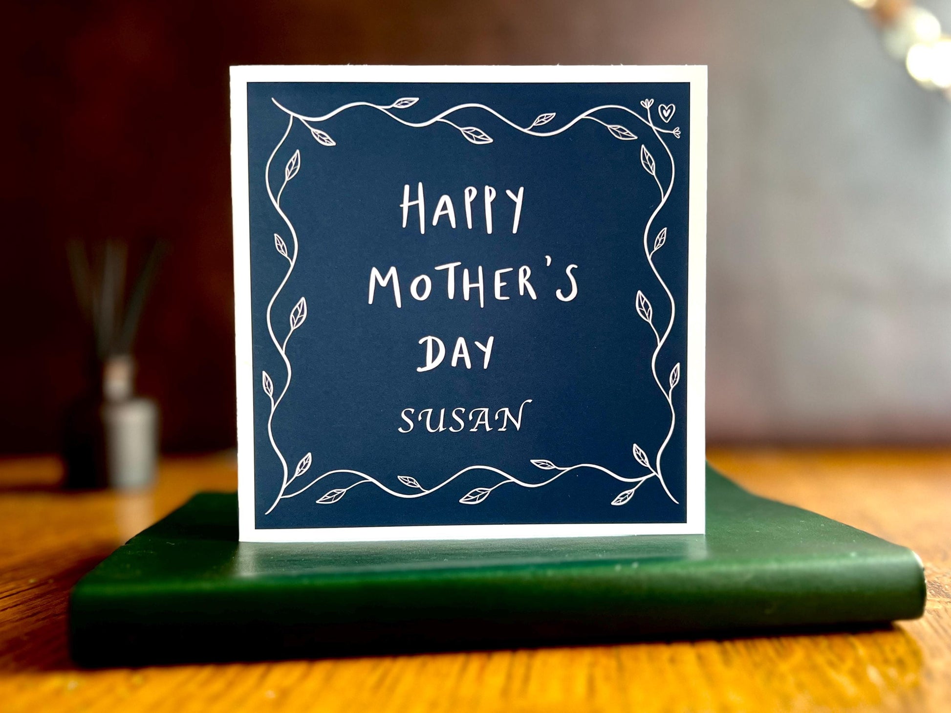 Artisan Mother’s Day greeting card with a dark navy blue background and simple leaf border, displayed standing upright on a tabletop. The personalised version shows “Happy Mother’s Day” with an added custom name underneath, such as Mum, grandma, or step-mum.
