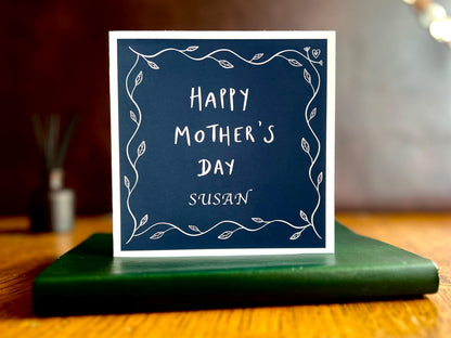 Artisan Mother’s Day greeting card with a dark navy blue background and simple leaf border, displayed standing upright on a tabletop. The personalised version shows “Happy Mother’s Day” with an added custom name underneath, such as Mum, grandma, or step-mum.