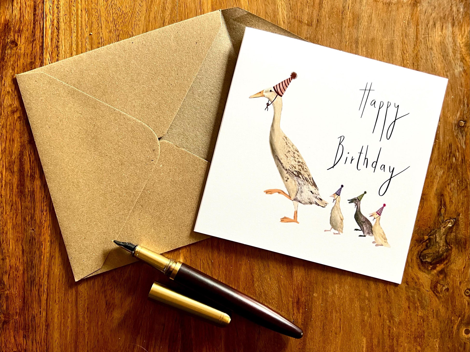 Artisan illustrated birthday greeting card featuring a mother runner duck with three ducklings wearing party hats, laid flat on a tabletop with a recycled fleck kraft envelope and pen. Playful countryside farm animal design on a white background with “Happy Birthday” text.