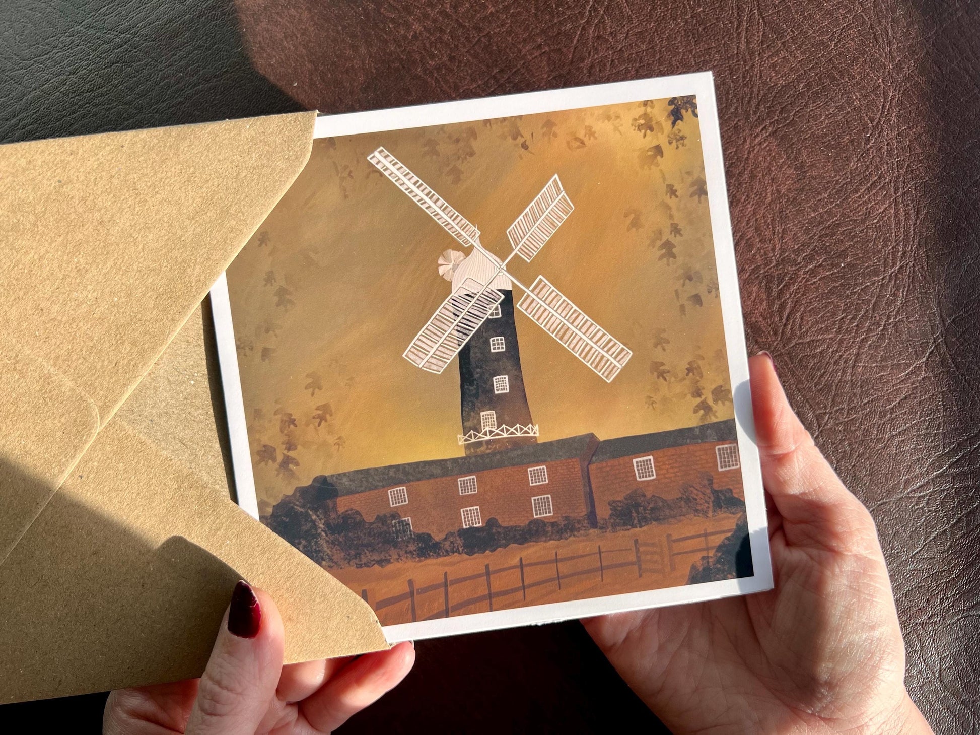 Artisan illustrated greeting card featuring Skidby Windmill in East Yorkshire standing behind farm buildings under an autumnal orange sunset sky. Countryside landscape art greeting card inspired by rural Yorkshire scenery.