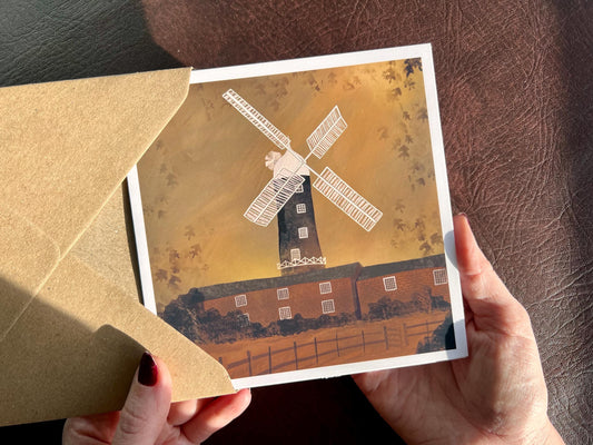 Artisan illustrated greeting card featuring Skidby Windmill in East Yorkshire standing behind farm buildings under an autumnal orange sunset sky. Countryside landscape art greeting card inspired by rural Yorkshire scenery.