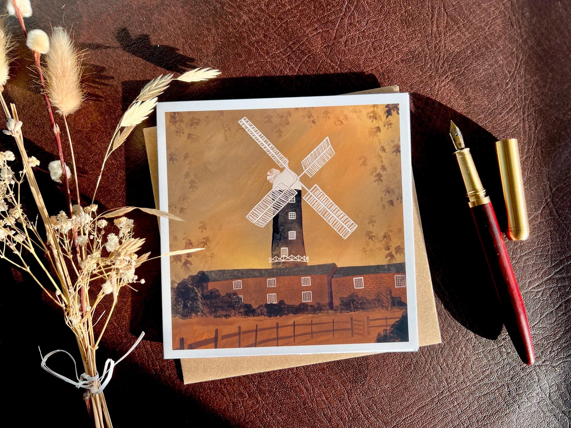 Artisan illustrated greeting card featuring Skidby Windmill in East Yorkshire behind farm buildings under an autumnal orange sunset sky, laid flat on a recycled fleck kraft envelope on a brown leatherette tabletop. Styled flat lay includes a fountain pen and dried flowers, showcasing countryside landscape art greeting card design.