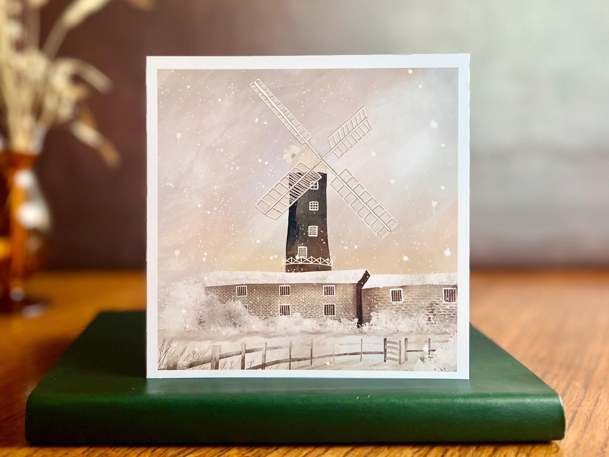 Artisan illustrated Christmas greeting card featuring Skidby Windmill in East Yorkshire behind snow-covered farm buildings under a pink and purple winter sunset sky with falling snow, displayed standing upright on a tabletop. Festive countryside winter landscape card design.
