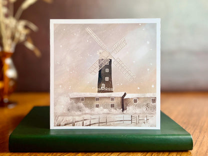 Artisan illustrated Christmas greeting card featuring Skidby Windmill in East Yorkshire behind snow-covered farm buildings under a pink and purple winter sunset sky with falling snow, displayed standing upright on a tabletop. Festive countryside winter landscape card design.