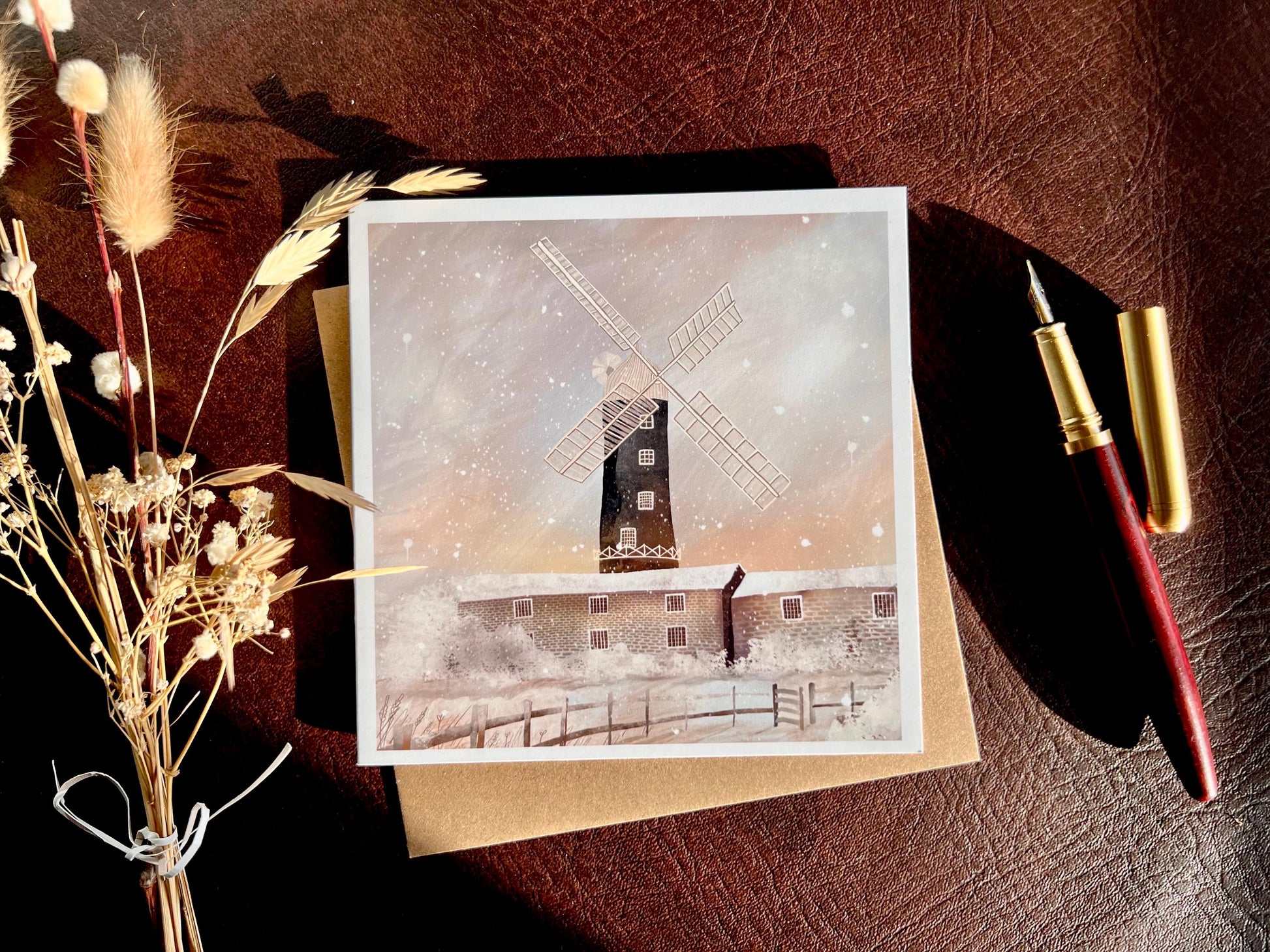 Artisan illustrated Christmas greeting card featuring Skidby Windmill in East Yorkshire behind snow-covered farm buildings under a pink and purple winter sunset sky with falling snow, styled in a sunlit flat lay on top of a recycled fleck kraft envelope. The scene includes a fountain pen and dried flowers on a leatherette surface. Festive countryside winter landscape greeting card design.
