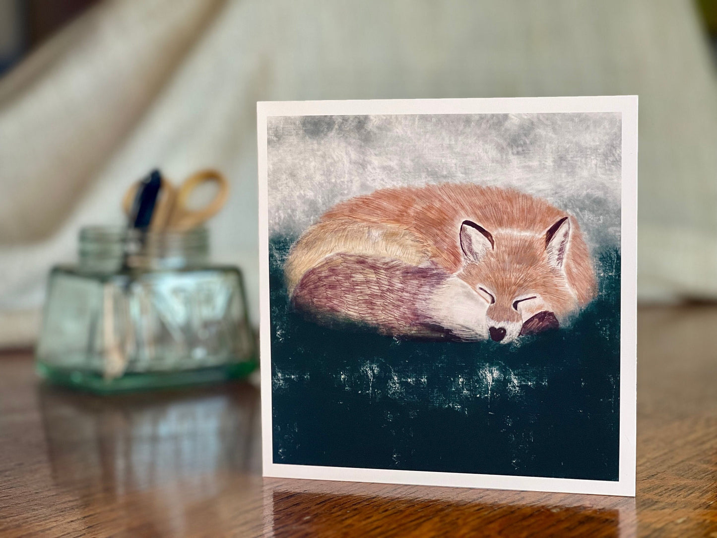 Artisan illustrated art greeting card featuring a sleeping fox in a calm woodland scene, set against a textured dark green and grey background, displayed standing upright on a tabletop. Versatile wildlife art card suitable for any occasion.
