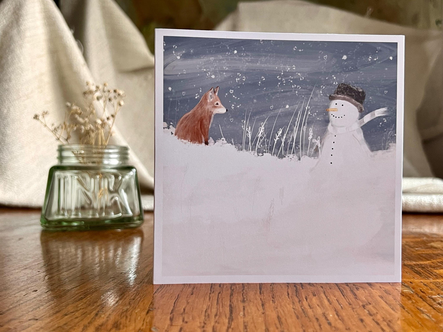 Artisan illustrated Christmas greeting card featuring a happy snowman wearing a hat looking at a fox cub in a snowy winter landscape, displayed standing upright on a tabletop. Festive wildlife-inspired seasonal greeting card design.