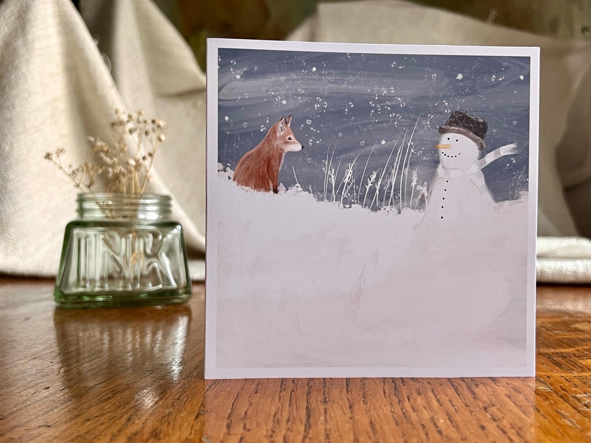 Artisan illustrated Christmas greeting card featuring a happy snowman wearing a hat looking at a fox cub in a snowy winter landscape, displayed standing upright on a tabletop. Festive wildlife-inspired seasonal greeting card design.