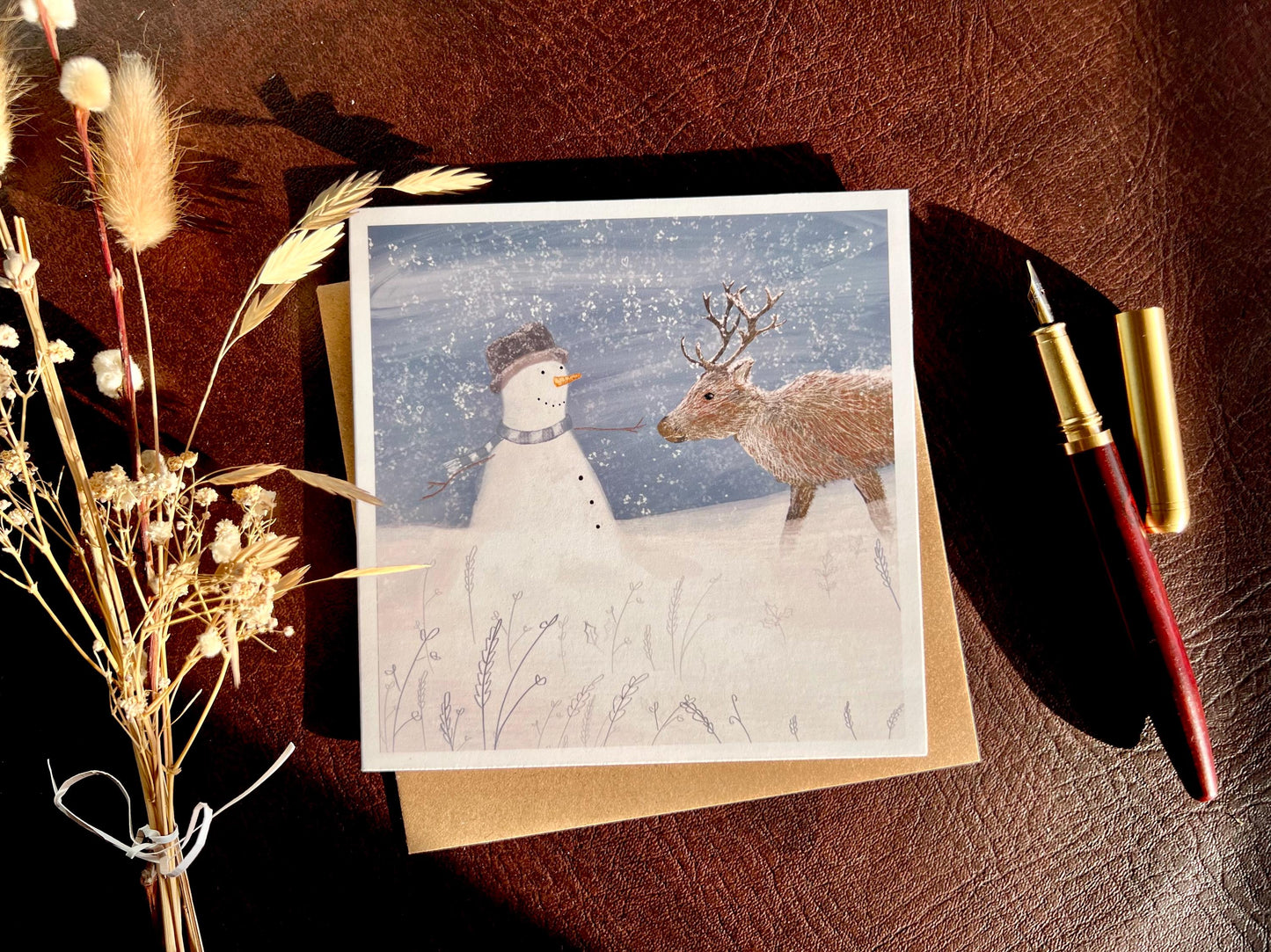 Artisan illustrated Christmas greeting card featuring a happy snowman wearing a hat looking at a reindeer in a snowy winter landscape, styled in a flat lay with a recycled fleck kraft envelope and dried flowers. The card is shown open to display the design, highlighting a festive wildlife-inspired seasonal greeting card scene.