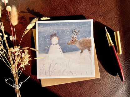 Artisan illustrated Christmas greeting card featuring a happy snowman wearing a hat looking at a reindeer in a snowy winter landscape, styled in a flat lay with a recycled fleck kraft envelope and dried flowers. The card is shown open to display the design, highlighting a festive wildlife-inspired seasonal greeting card scene.