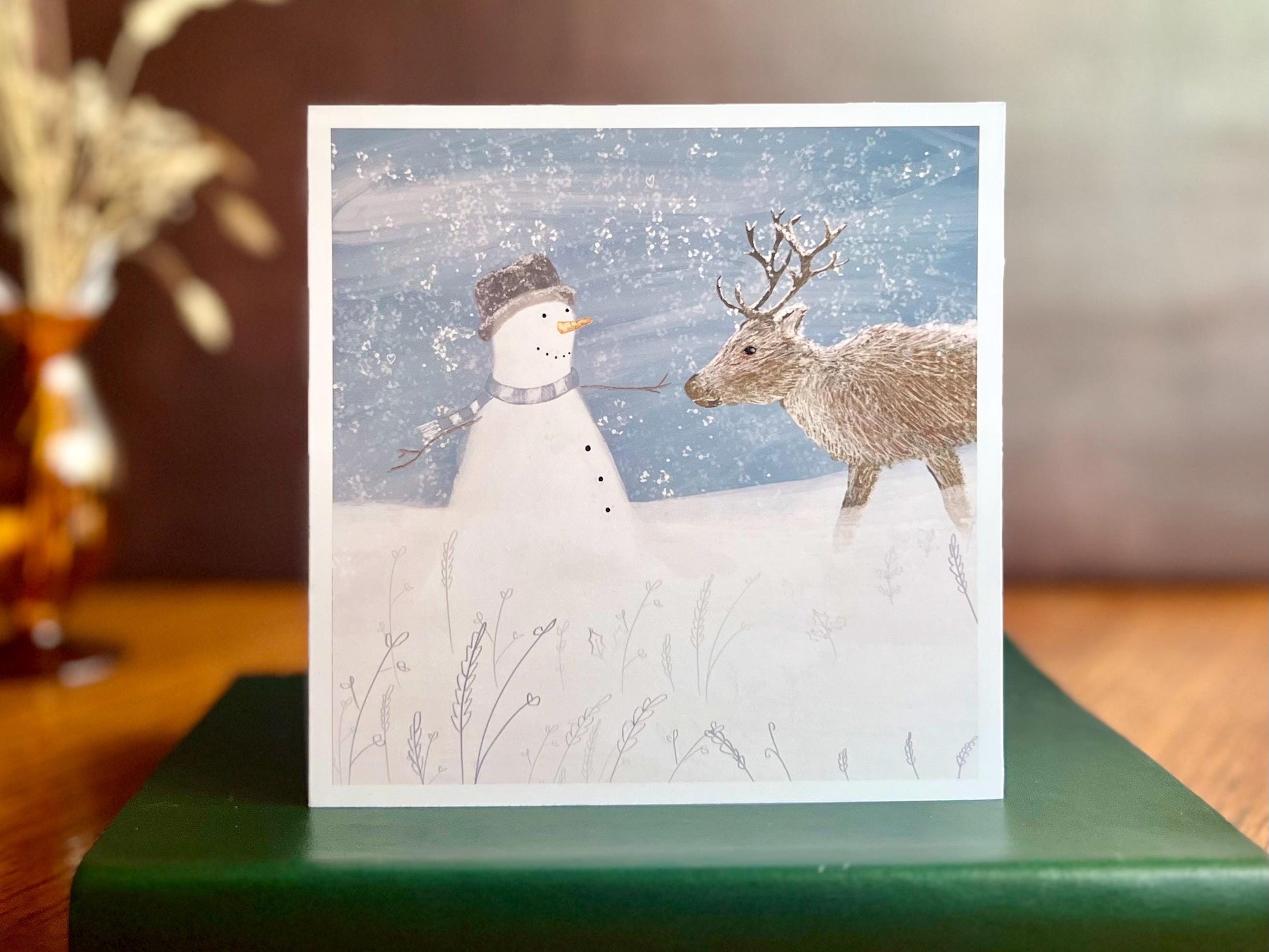 Artisan illustrated Christmas greeting card featuring a happy snowman wearing a hat looking at a reindeer in a snowy winter landscape, displayed standing on a green leatherette book. Festive wildlife-inspired seasonal greeting card design.