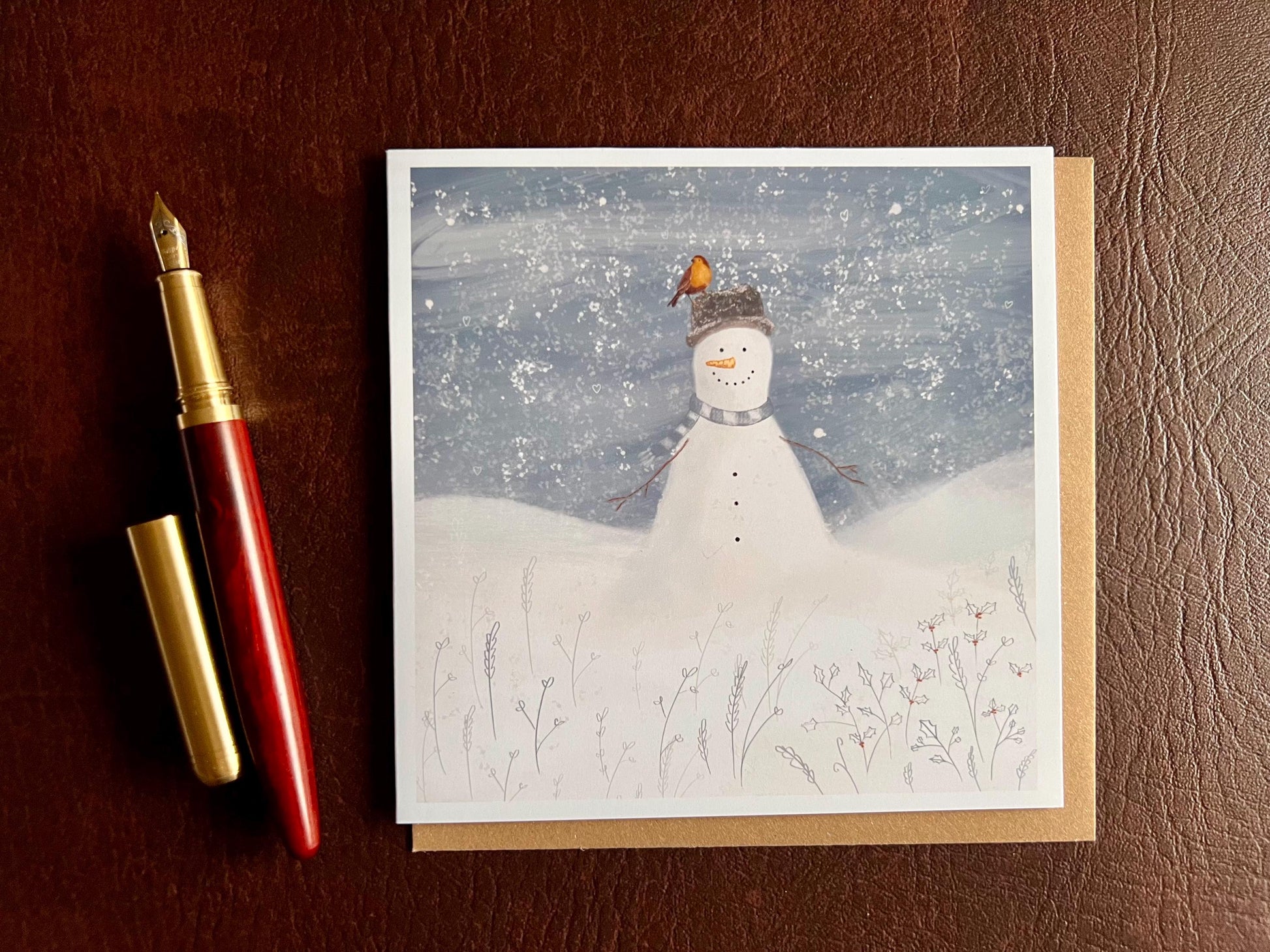Artisan illustrated Christmas greeting card featuring a happy snowman wearing a hat with a robin perched on top, set in a cheerful snowy winter landscape, laid flat on a leatherette tabletop. Festive wildlife-inspired seasonal greeting card design.