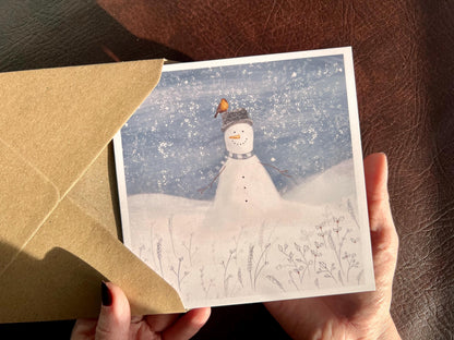 Artisan illustrated Christmas greeting card featuring a happy snowman wearing a hat with a robin perched on top, set in a cheerful snowy winter landscape. Festive wildlife-inspired seasonal greeting card design.