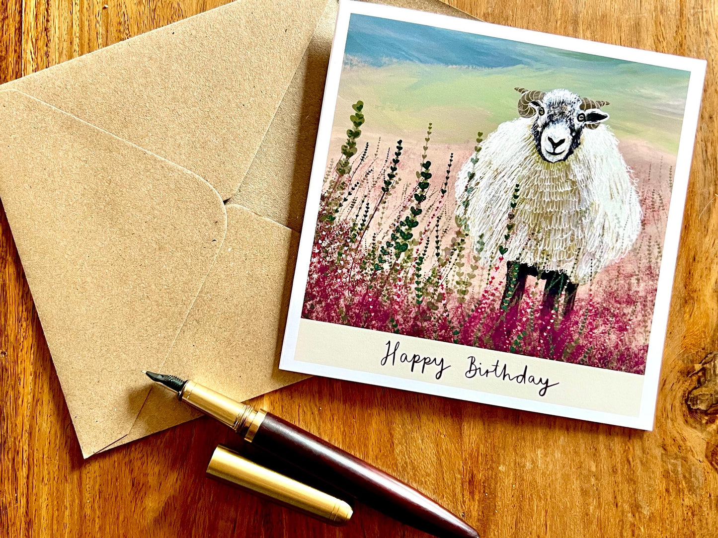 Artisan illustrated birthday greeting card featuring a Swaledale sheep standing in a heather landscape on the Yorkshire moors, laid flat on a wooden tabletop with a recycled fleck kraft envelope and pen. Countryside wildlife art card design with “Happy Birthday” text beneath the illustration.