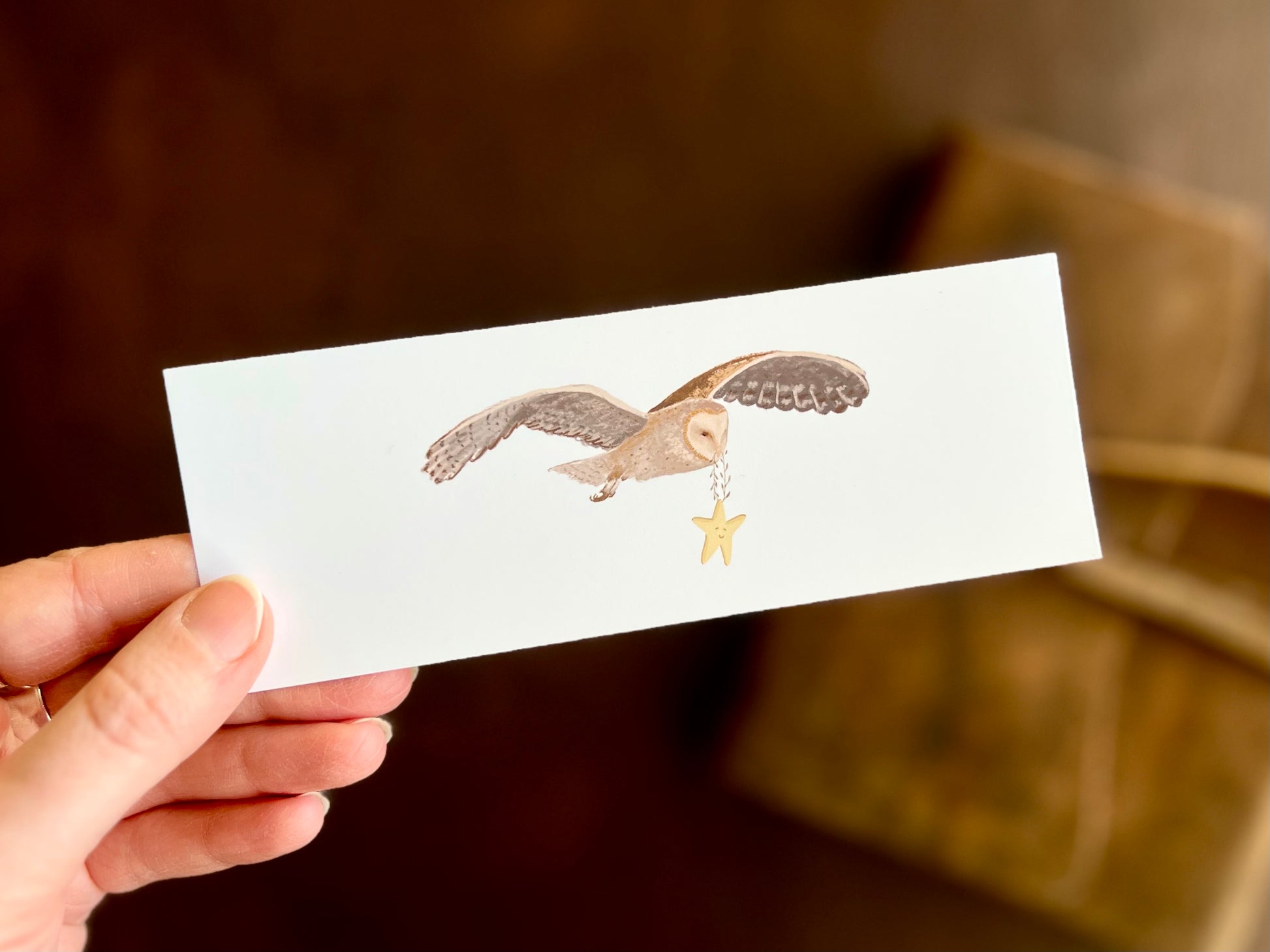 Hand holding a white bookmark with illustrated barn owl design. The magical owl is in fight, carrying a smiling star. Whimsical illustration by artist Hayley Victoria. 