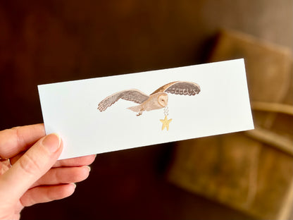 Hand holding a white bookmark with illustrated barn owl design. The magical owl is in fight, carrying a smiling star. Whimsical illustration by artist Hayley Victoria. 