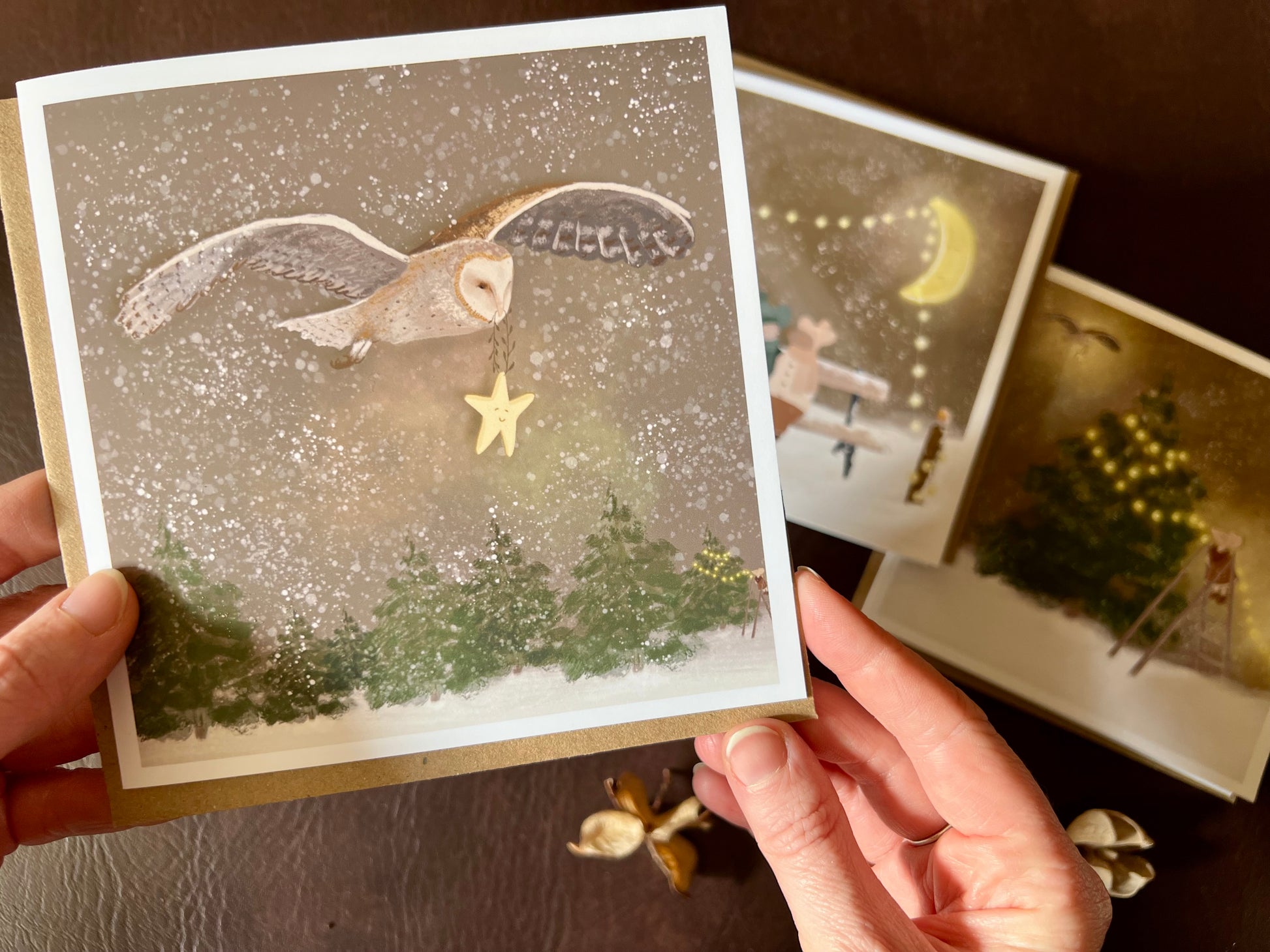 Premium Christmas card from a set of three woodland designs, featuring a barn owl flying through a snowy night sky carrying a smiling star. The storybook-style winter scene is shown in close-up being held in two hands, with the other two designs softly out of focus in the background. Ideal festive greeting card for animal lovers and rustic Christmas card sets. Printed on thick 312gsm fine art card by a UK independent artist.