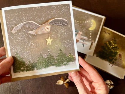 Premium Christmas card from a set of three woodland designs, featuring a barn owl flying through a snowy night sky carrying a smiling star. The storybook-style winter scene is shown in close-up being held in two hands, with the other two designs softly out of focus in the background. Ideal festive greeting card for animal lovers and rustic Christmas card sets. Printed on thick 312gsm fine art card by a UK independent artist.