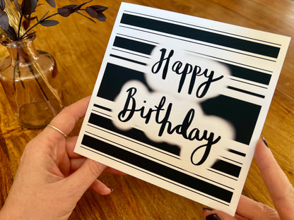 Aerial view of hands holding a handmade monochrome birthday card featuring a black and white hooped background with 'Happy Birthday' in cursive script, shown being examined in a lifestyle setting.