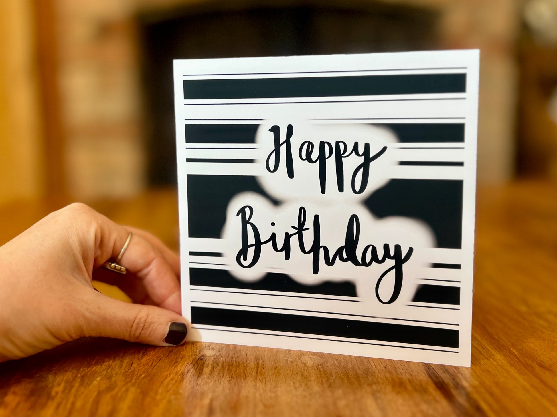 Hand placing a handmade monochrome birthday card upright onto a wooden tabletop. The card features a black and white hooped background with 'Happy Birthday' in cursive lettering, shown in a lifestyle setting.