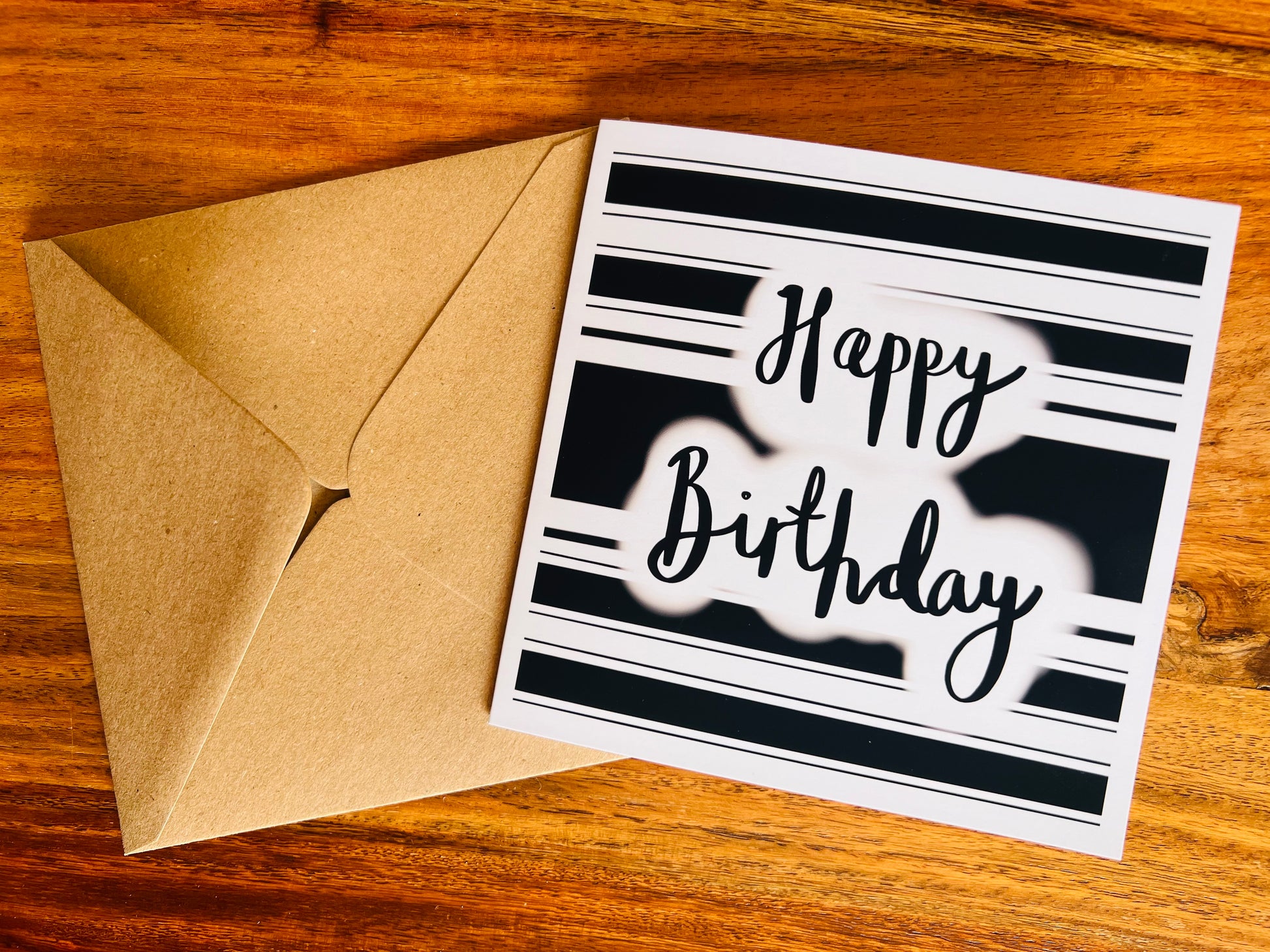 Flat lay image of a handmade monochrome birthday card featuring a black and white hooped background and 'Happy Birthday' in cursive text, shown laid on a tabletop with the kraft envelope alongside it.